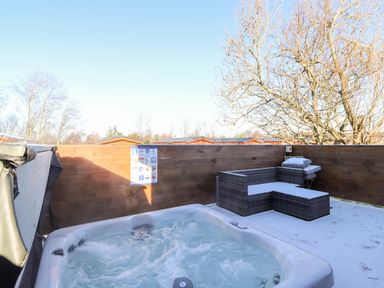 An outdoor hot tub with a wicker sectional sofa and cushions on a patio with wooden fencing and leafless trees at Orion in Felmoor Park