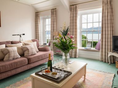 A living room with a pink sofa and pillows a white coffee table with a flower vase a tray with drinks and snacks and windows with views of water at Tros Yr Afon in Beaumaris