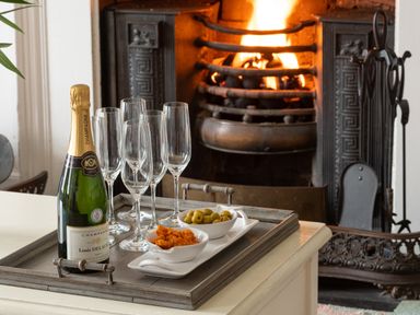 A tray with a bottle of champagne glasses and bowls of snacks near a lit fireplace at Tros Yr Afon in Beaumaris