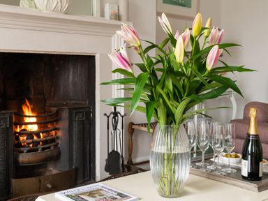 A vase with pink lilies on a table next to a fireplace with a fire burning at Tros Yr Afon in Beaumaris