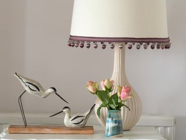 A table lamp with a fabric shade next to a small vase with pink roses and a wooden bird sculpture at Tros Yr Afon in Beaumaris