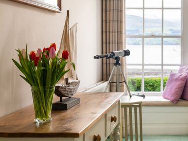 A vase with tulips on a wooden table with a model sailboat and a telescope by a window with cushions and curtains at Tros Yr Afon in Beaumaris