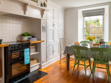 A kitchen with a wooden floor a black oven a white cupboard and a table with green chairs by a window at Tros Yr Afon in Beaumaris