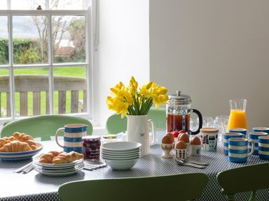 A dining table set with croissants jam coffee cups eggs orange juice and a vase of yellow flowers near a window at Tros Yr Afon in Beaumaris