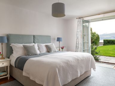 A bedroom with a large bed a blue headboard bedside tables with lamps an open door to a garden at Tros Yr Afon in Beaumaris