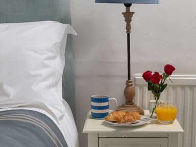A bedside table with a lamp, striped mug, plate with croissants and knife, glass of orange juice, and small vase with red roses next to a bed at Tros Yr Afon in Beaumaris