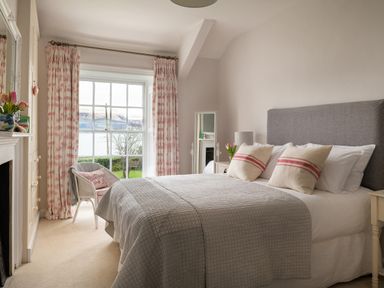 A bedroom with a bed, pillows, chair, curtains, and a window with a view of water at Tros Yr Afon in Beaumaris