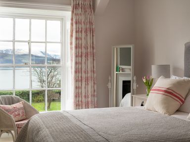 A bedroom with a large window showing a lake and mountains with a bed a pillow a bedside table lamp flowers a mirror and a wicker chair at Tros Yr Afon Beaumaris