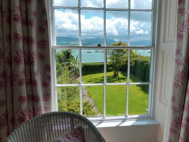 A view of a garden with a lawn tree and hedges and water with boats in the distance seen through a multi pane window with patterned curtains at Tros Yr Afon in Beaumaris