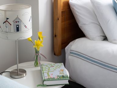 A bedside table with a lamp yellow flowers in a glass vase and a book next to a bed with white sheet and blue stripes at Tros Yr Afon in Beaumaris