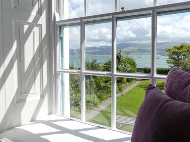 A window seat with purple cushions overlooking a garden path leading to a lake with hills in the background at Tros Yr Afon in Beaumaris