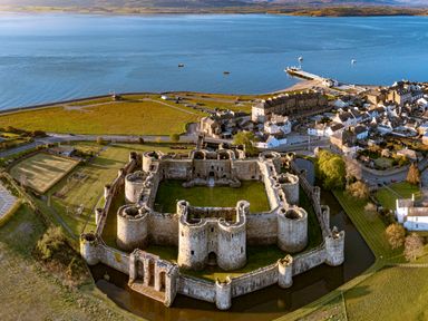 An aerial view of a large stone castle with circular towers surrounded by a moat near a coastal town with houses and a pier at Tros Yr Afon in Beaumaris