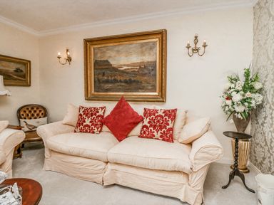 A living room with a beige sofa with red pillows a framed painting on the wall candles a flower arrangement and a brown chair at Storrs Lodge in Bowness-On-Windermere