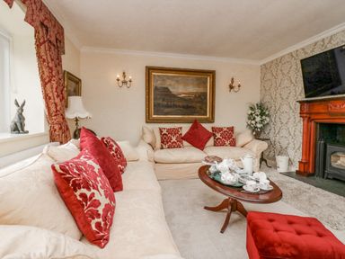 A living room with beige sofas and red cushions a wooden coffee table with tea set a red ottoman a fireplace and a painting at Storrs Lodge in Bowness-On-Windermere