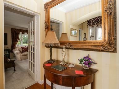 A hallway table with a lamp statue flowers and books below a large ornate mirror at Storrs Lodge in Bowness-On-Windermere