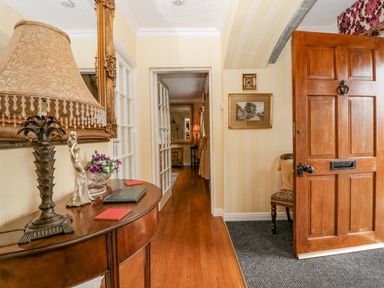An entryway with a wooden door, a small table with a lamp, decorative figurine, and flowers, leading to a hallway with wooden floors and paintings on the walls at Storrs Lodge in Bowness-On-Windermere