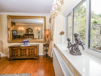 A room with a large mirror above a wooden cabinet a table lamp on a side table and sculptures on a windowsill at Storrs Lodge in Bowness-On-Windermere