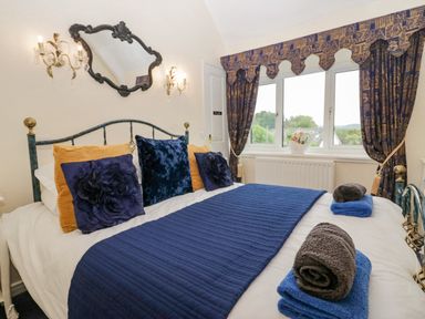 A bedroom with a bed covered in blue and yellow pillows and blue bed runner with rolled towels on top and a window with patterned curtains at Storrs Lodge in Bowness-On-Windermere