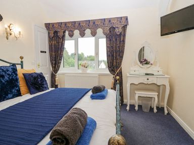 A bedroom with a bed and blue bedding a white dressing table with mirror and a wall-mounted television at Storrs Lodge in Bowness-On-Windermere