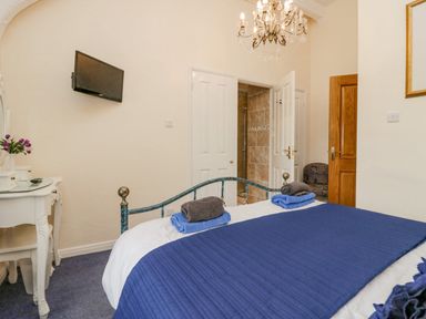 A bedroom with a bed covered with blue bedding and towels on it a white dressing table with a mirror and flowers a wall-mounted television two open doors showing a tiled bathroom and a wooden door at Storrs Lodge in Bowness-On-Windermere
