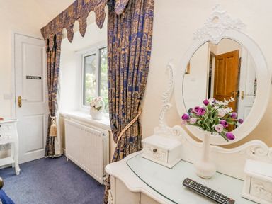 A dressing table with a round mirror and flowers in a vase next to a window with patterned curtains at Storrs Lodge in Bowness-On-Windermere