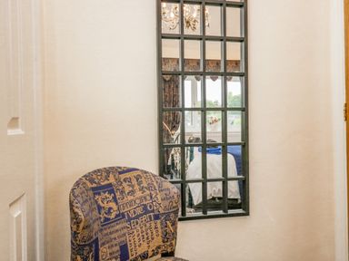 A patterned chair in front of a wall mirror reflecting a bedroom window with curtains and a bed with folded towels at Storrs Lodge in Bowness-On-Windermere