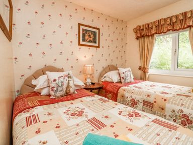 A bedroom with two single beds with floral bedspreads and bunny pillows a lamp on a bedside table and a window with curtains at Storrs Lodge in Bowness-On-Windermere