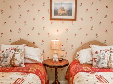 A bedroom with two single beds with red and beige bedding and rabbit pillows and a wooden table with a lamp between the beds at Storrs Lodge Bowness-On-Windermere