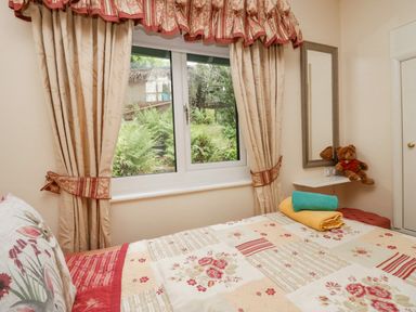 A bedroom with floral bedspread and folded towels on bed next to window with curtains and teddy bear on wall shelf at Storrs Lodge in Bowness-On-Windermere