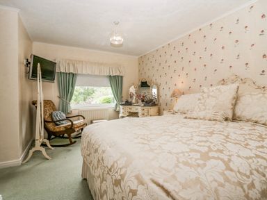 A bedroom with a large bed, floral wallpaper, a rocking chair, a dressing table with mirrors, and a window with green curtains at Storrs Lodge in Bowness-On-Windermere