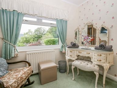 A vintage dressing table with mirror and stool next to a window with green curtains and a floral armchair at Storrs Lodge in Bowness-On-Windermere