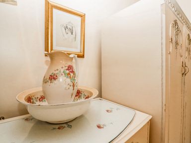 A ceramic pitcher and bowl with floral designs on a table next to a decorated cupboard and framed art at Storrs Lodge Bowness-On-Windermere