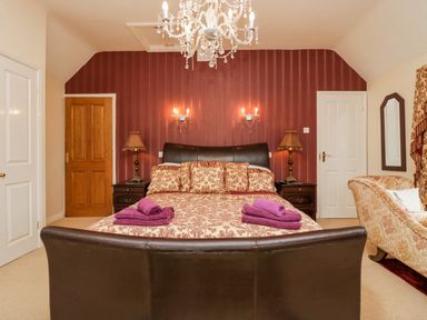 A bedroom with a double bed with patterned bedspread and purple towels a chandelier two bedside lamps and an armchair at Storrs Lodge in Bowness-On-Windermere