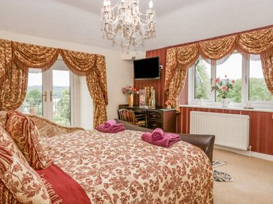 A bedroom with a bed with patterned bedspread and pillows a chandelier a desk with a chair and a television on the wall at Storrs Lodge in Bowness-On-Windermere