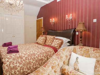 A bedroom with a patterned bedspread pillows and folded towels a patterned armchair with a rabbit pillow and lamps on a red striped wall at Storrs Lodge in Bowness-On-Windermere