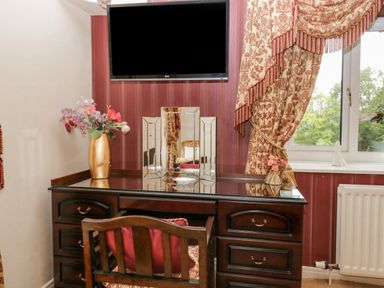 A wooden desk with drawers and a chair in front of a window with patterned curtains and a wall mounted television at Storrs Lodge in Bowness-On-Windermere
