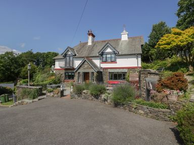 A two-story house with stone and white walls surrounded by greenery and a paved driveway at Storrs Lodge in Bowness-On-Windermere
