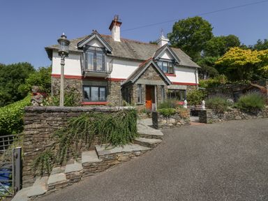 A stone and white house with a sloped roof a small balcony a wooden door and stone steps at Storrs Lodge in Bowness-On-Windermere