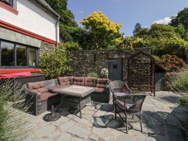 An outdoor patio with wicker furniture including a corner sofa and chairs a table stone flooring and a stone wall with a door at Storrs Lodge in Bowness-On-Windermere