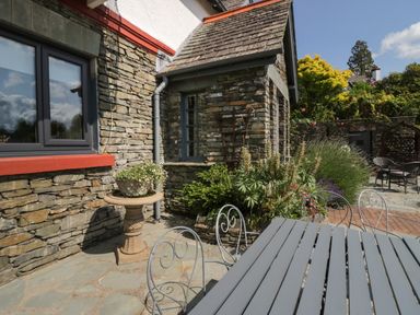 An outdoor stone patio area with a metal table and chairs next to a stone house with bushes and a flower pot on a pedestal at Storrs Lodge in Bowness-On-Windermere