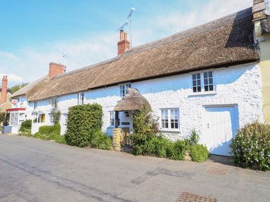 A long white thatched roof cottage with windows and a door surrounded by bushes and greenery at Lime Cottage in Burton Bradstock