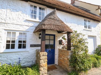 The entrance of a white stone cottage with a blue door and thatched porch at Lime Cottage in Burton Bradstock