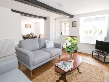 A living room with grey sofas a wooden coffee table with flowers in a vase and magazines a dining area visible through an open doorway at Lime Cottage in Burton Bradstock