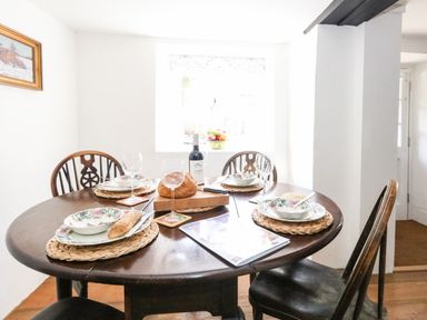 A dining room with a round wooden table set for four with plates bowls wine glasses bread and a bottle of wine at Lime Cottage in Burton Bradstock