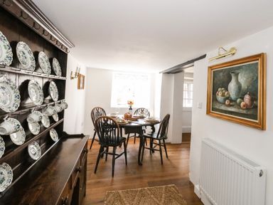 A dining room with wooden chairs and table set near a window alongside a shelf displaying plates and cups at Lime Cottage in Burton Bradstock