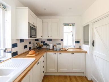 A kitchen with white cabinets wooden countertops a microwave a bread loaf a wine rack and a white door at Lime Cottage in Burton Bradstock