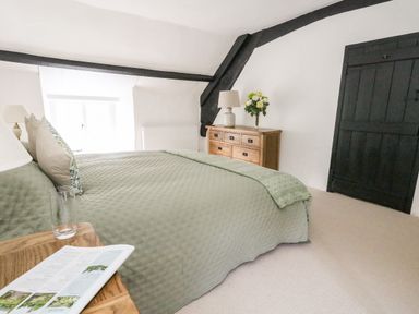 A bedroom with a bed covered in green bedding a wooden dresser with a lamp and flowers and a black wooden door at Lime Cottage in Burton Bradstock