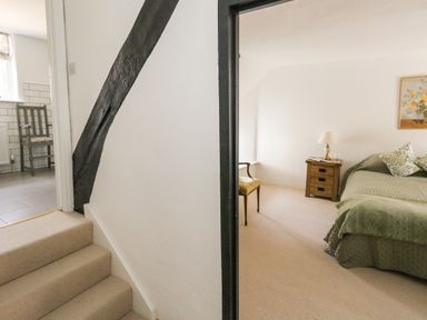 A bedroom with bed side table lamp and chair next to stairs leading to a kitchen with chair at Lime Cottage Burton Bradstock