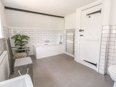 A bathroom with a bathtub a towel rack a wooden chair a plant and a white door at Lime Cottage in Burton Bradstock