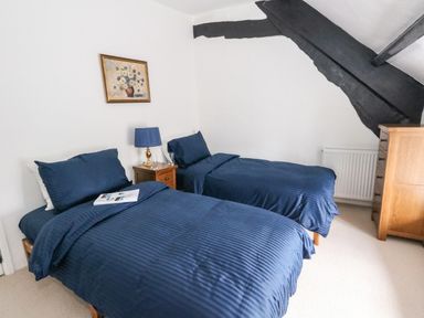 A bedroom with two single beds with blue bedding a wooden nightstand with a lamp and a wooden dresser at Lime Cottage in Burton Bradstock